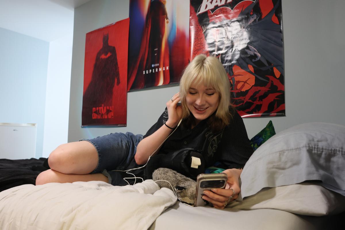 A student lays on their bed in their residence room, looking at their phone, with posters on the wall behind them.
