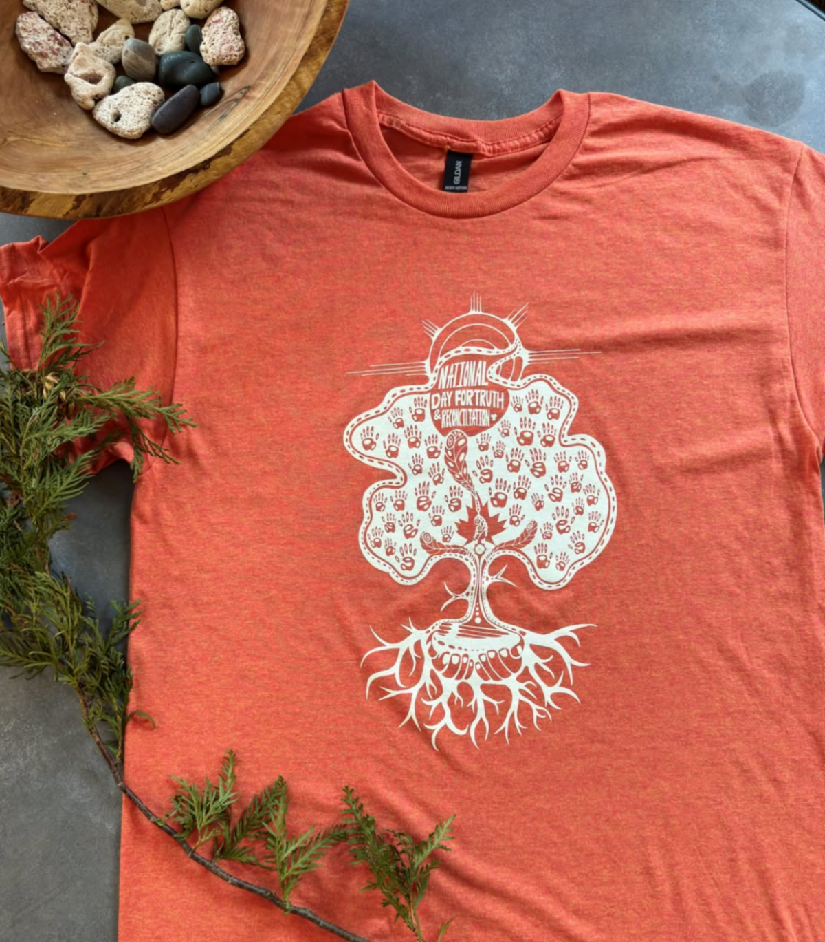An orange shirt with the artwork, "Rooted Remembrance" by Mazinibii’ige, lies flat with medicines around it.