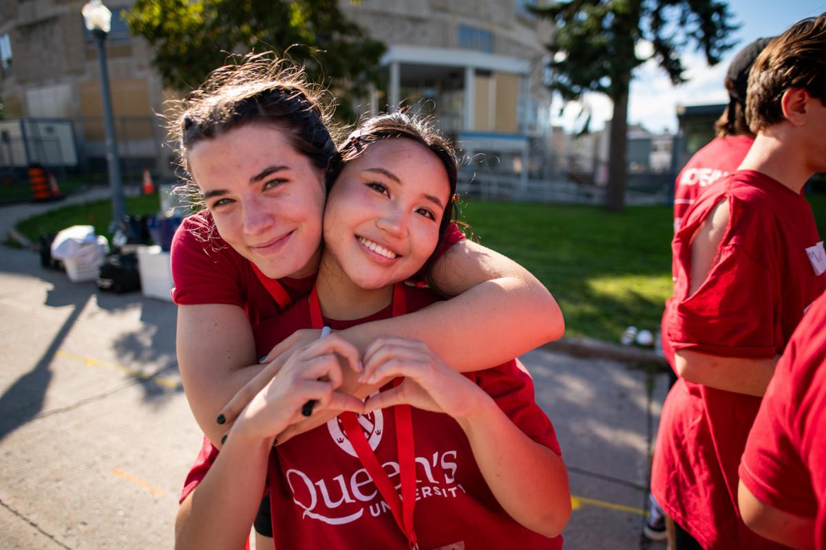 Two students smile, the first with their arms around the other, and the second makes a heart with their hands.