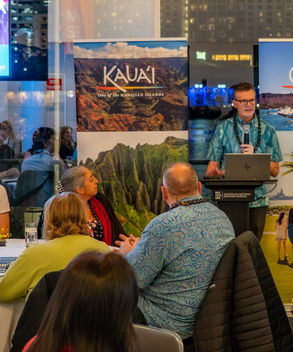 A presenter stands at a podium with a laptop, speaking to an audience seated at tables. Behind him are banners for "Kauai" and "Oahu", both described as "One of the Hawaiian Islands".