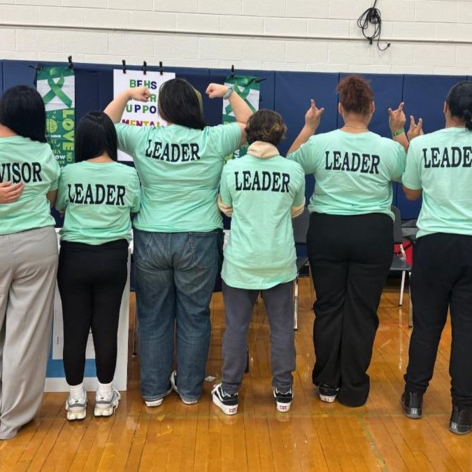 Group of teens stand together in a row with backs to camera wearing matching T-shirts reading "Leader" on the back