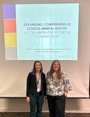 Rebekah and Rosie pose in front of the projector screen at their presentation. A slide on the screen behind them reads, "Expanding Comprehensive School Mental health: A Collaborative Effort in Connecticut."