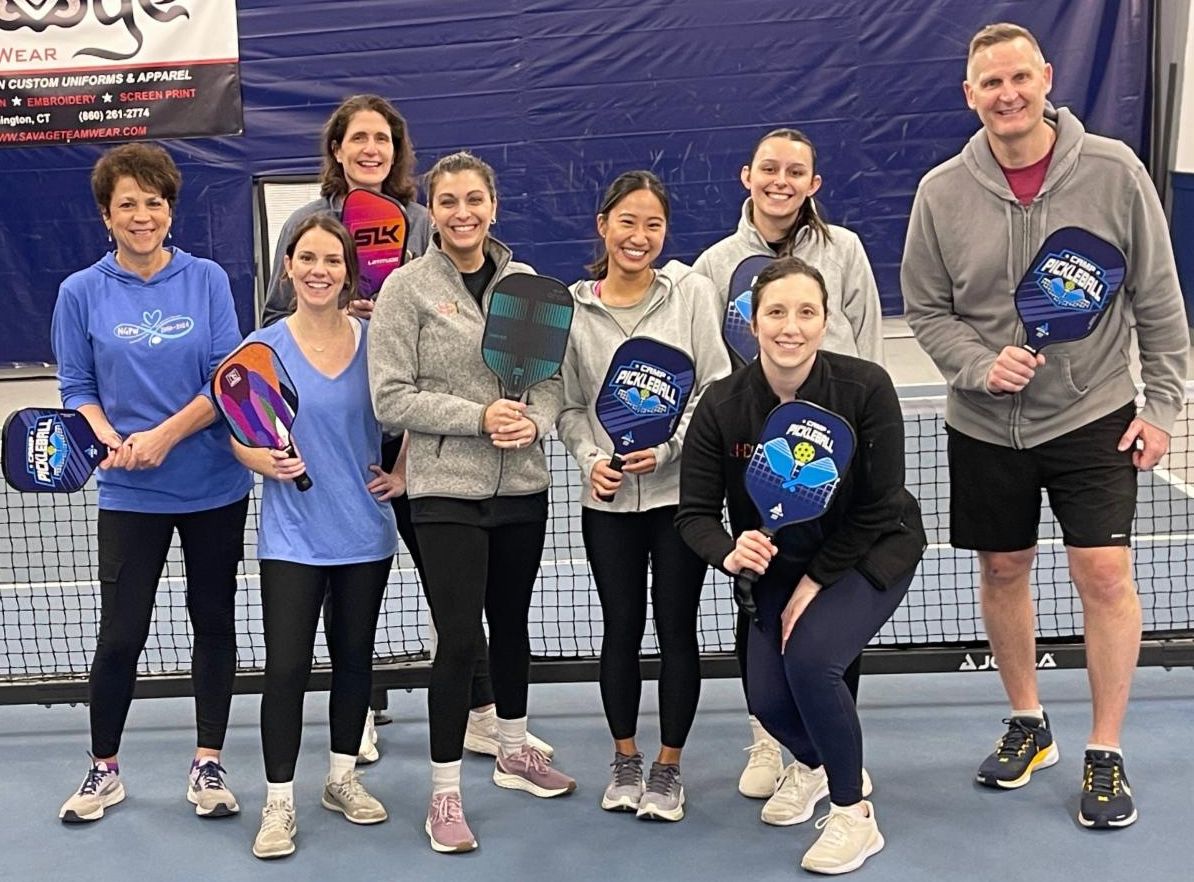 A group of eight CHDI staff stand together with pickleball paddles on pickleball court.