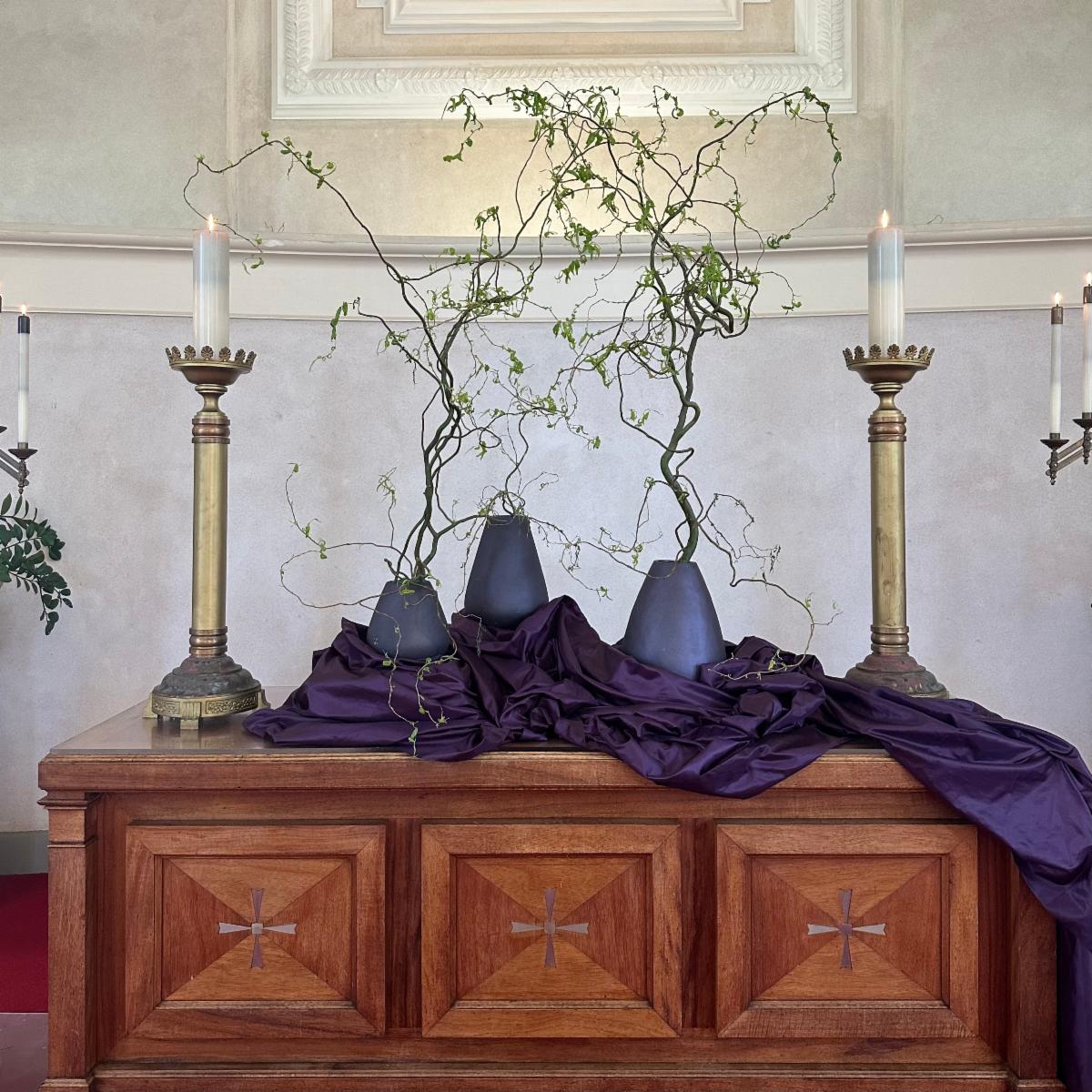 Altarscape on first Sunday of Lent, three purple vases with thin winding branches and sparse leaves, purple draped cloth on table