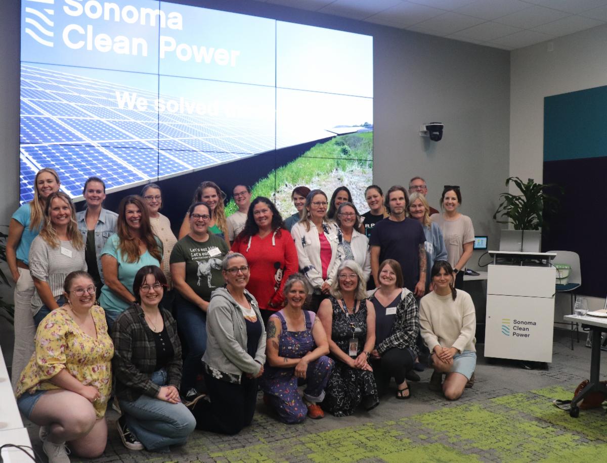 a group of approx 20 teachers some kneeling some standing in front of a large video screen posing for a group photo
