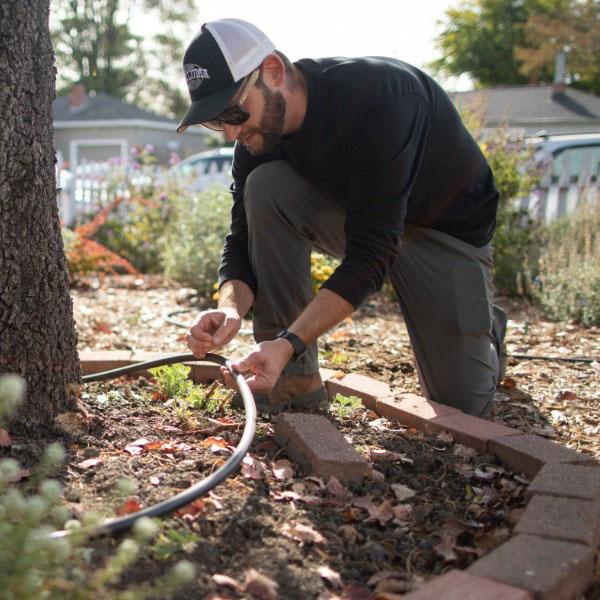 a man with a baseball cap and sunglasses crouches down to adjust a drip irrigation line in the landscaping