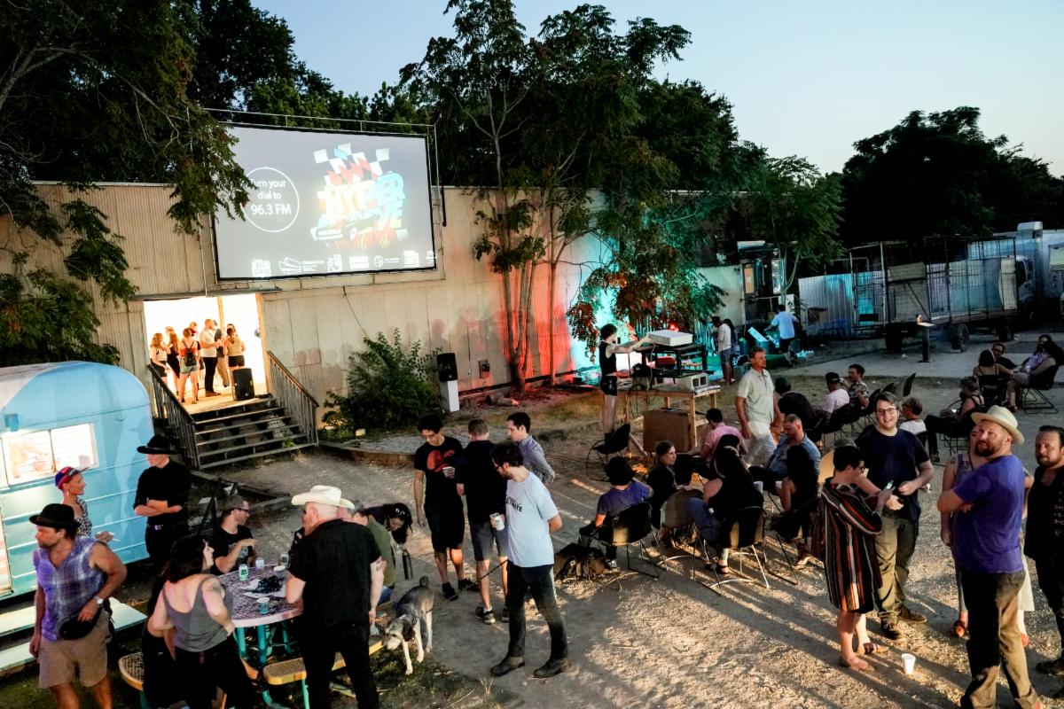 People stand outside a venue in the dusk while a crowd can be seen inside.