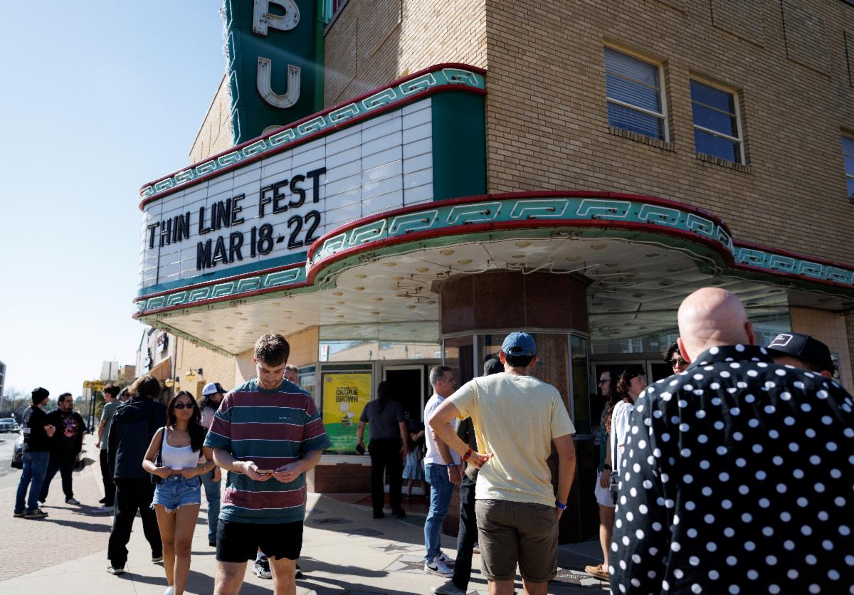People stand outside a classic theater, featuring a marque that reads, "Thin Line Fest Mar 18-22."