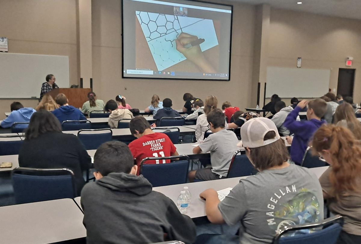 A classroom of high school-aged students is seen sitting at tables facing a projection screen while an artist demonstrates an art technique.