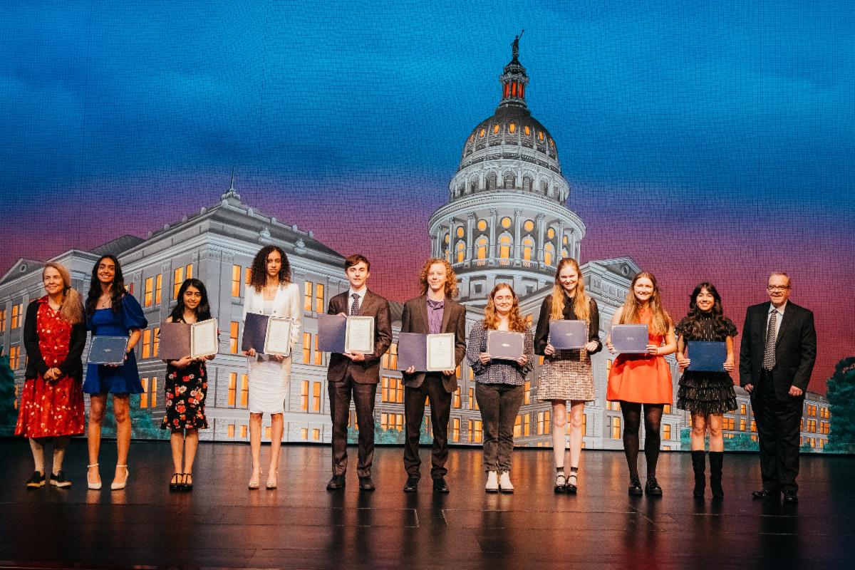 Nine students stand on a stage holding certificates. Naomi Shihab Nye stands on the left and Gary Gibbs stands on the right. A stylized image of the Texas Capitol dome is behind them.