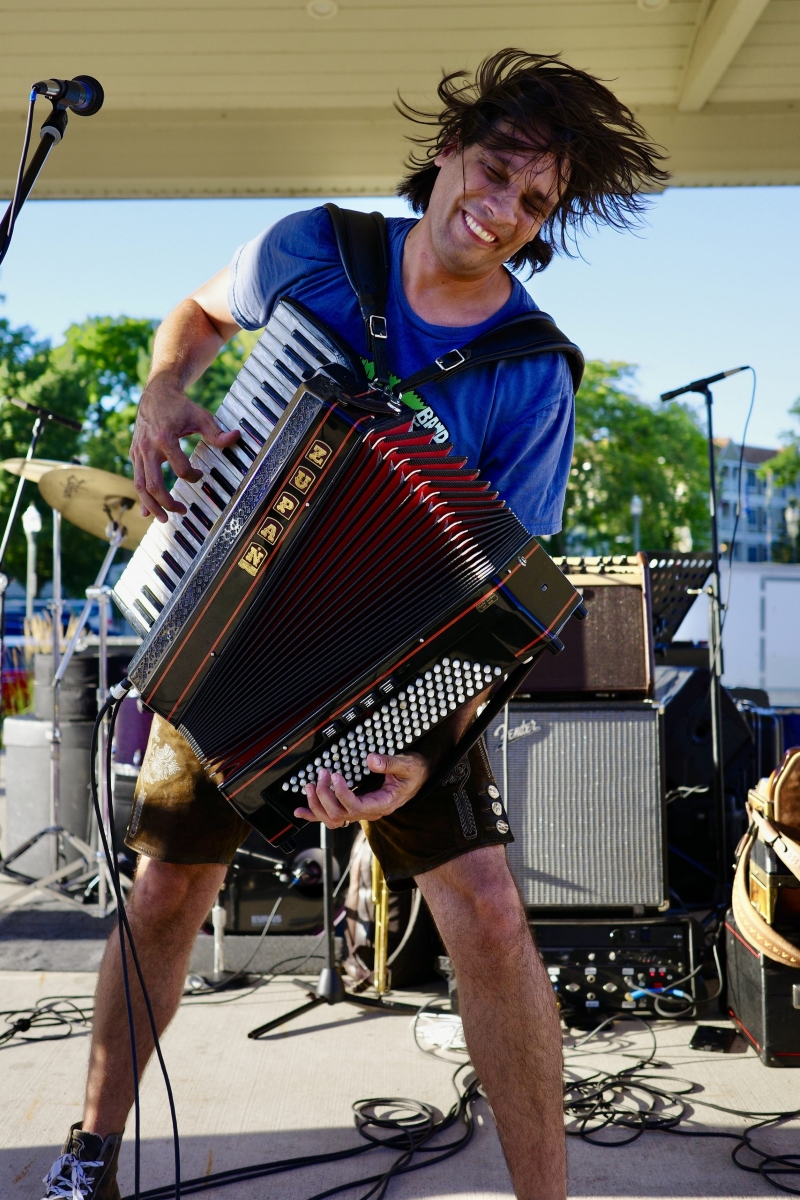 Alex Meixner smiles and plays the accordion while standing on an outdoor stage.