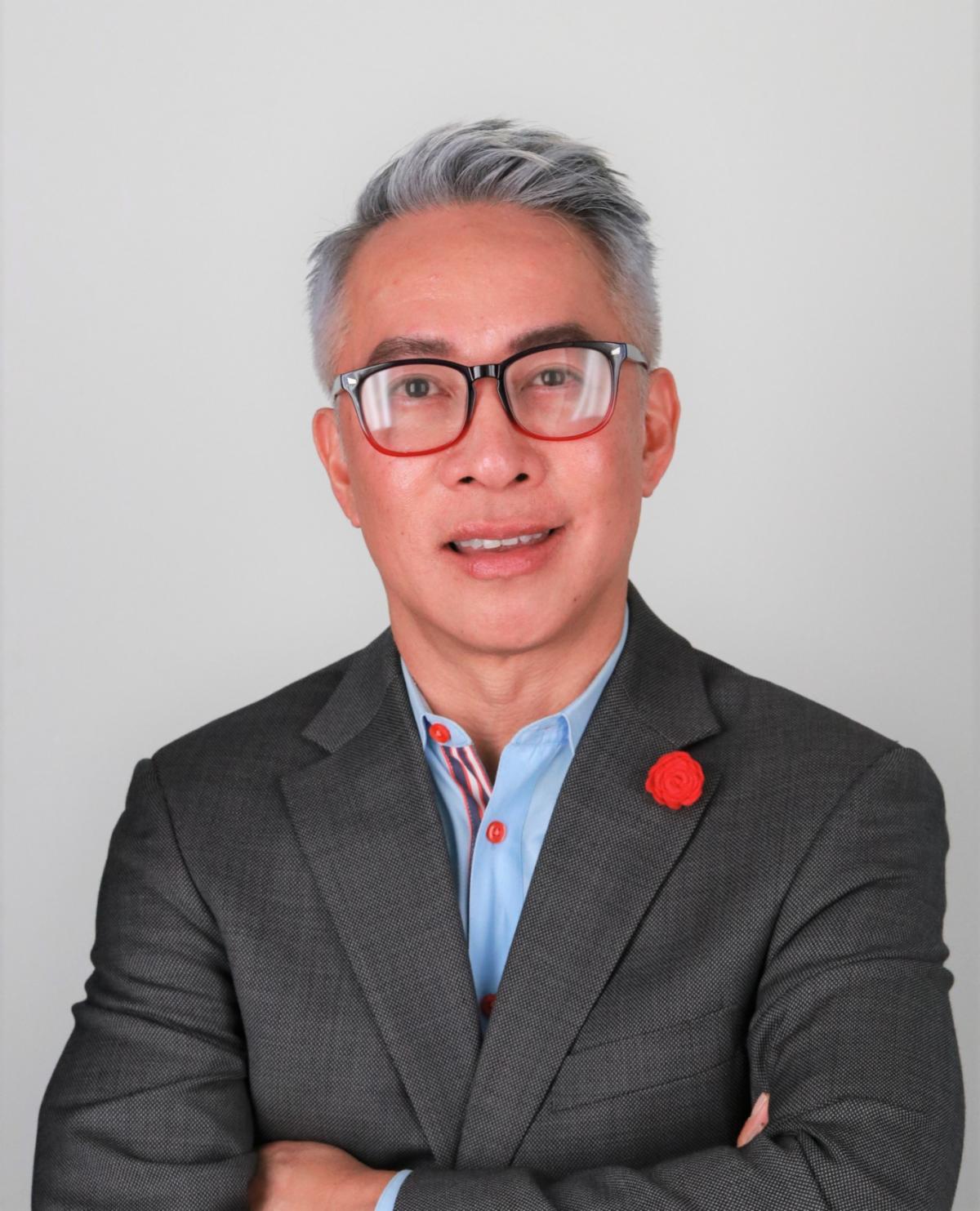 Bao-Long Chu is shown from the chest up with his arms crossed in front of him. He wears glasses and smiles. His greying dark hair is swept upward. He wears a grey jacket and blue button-up.