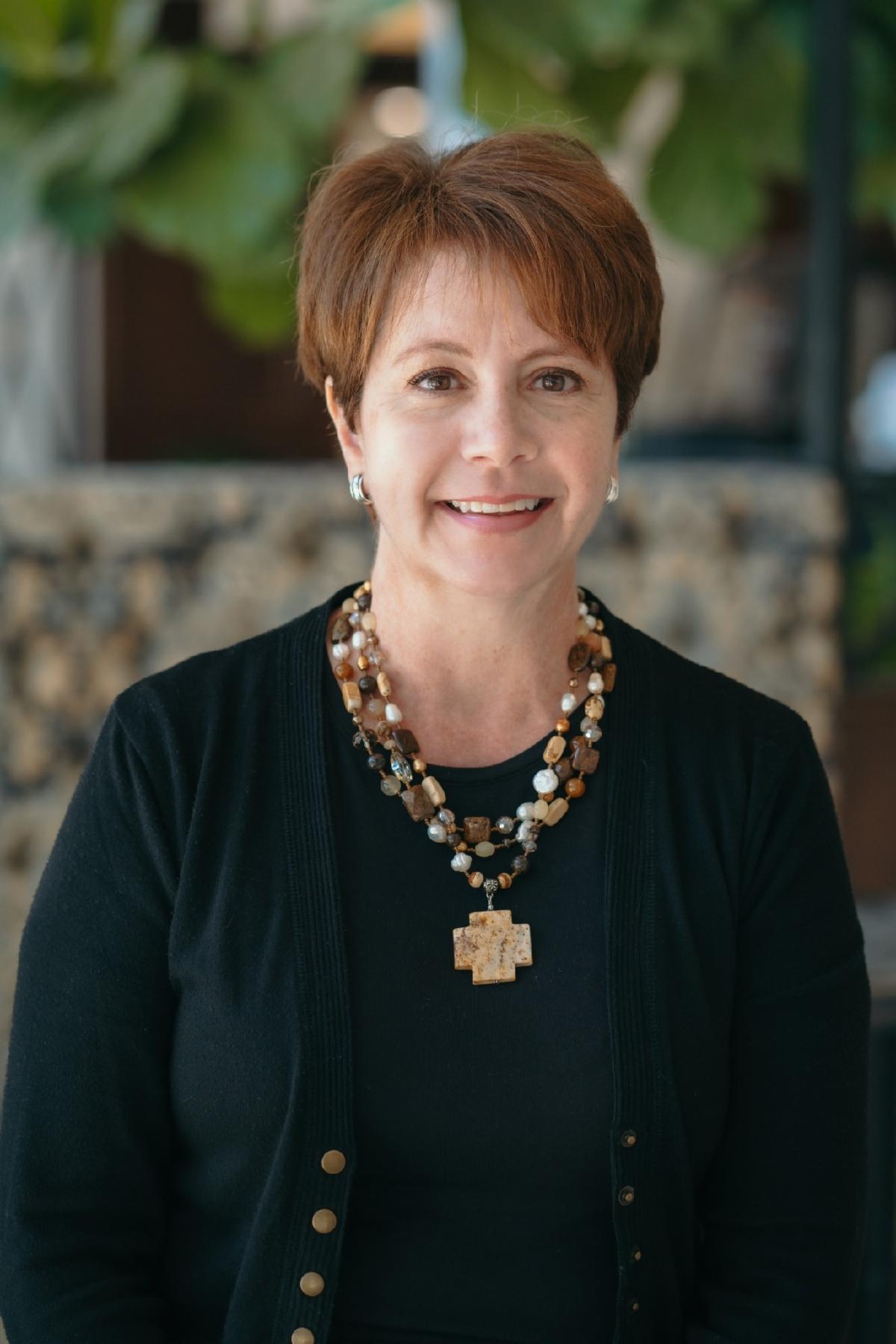 Stacy Keith smiles at the camera. She has short, dark red hair. She wears a dark shirt and a gold necklace.