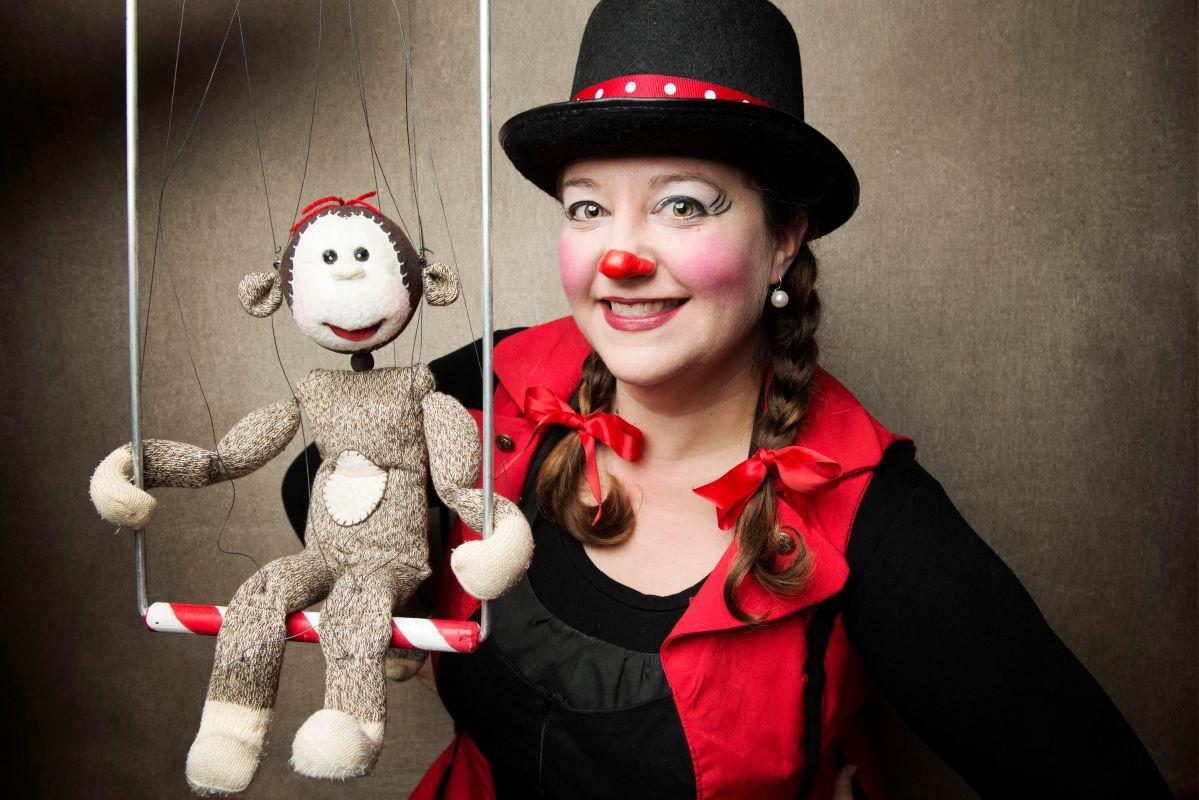 A woman in light clown makeup and a bowler hat poses with a sock monkey puppet.