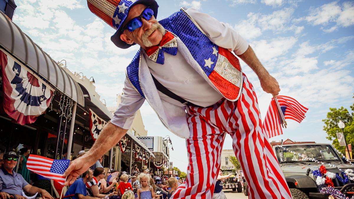 A man in patriotic garb holding American flags leans over the camera along a parade route.