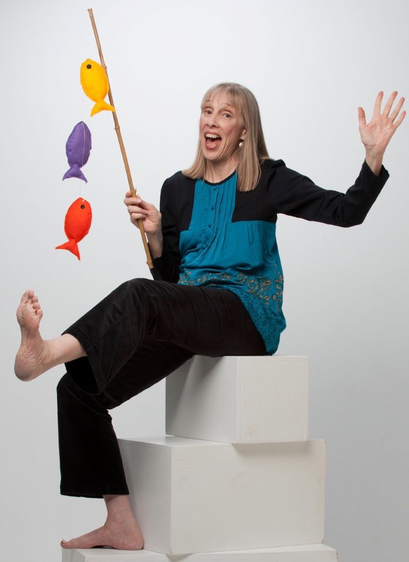Sue Young sits on two white blocks and holds a string of colorful cloth fish. She waves her other hand and kicks a bare foot out.
