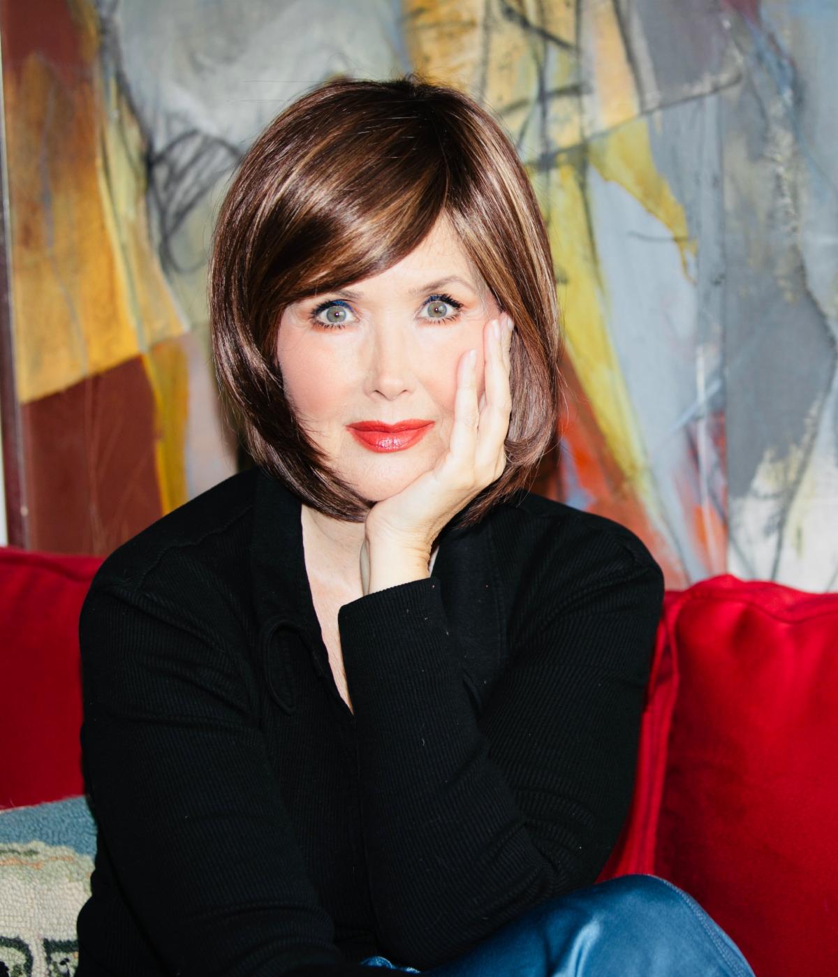 Janine Turner looks at the camera with her left hand cupping the side of her face. She has a dark brown bob with sideswept bangs and wears a dark shirt.
