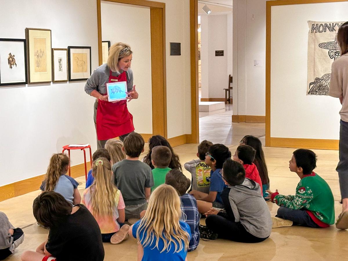 A woman in a red apron holds up an image of an art work for children, seated in a gallery, to see.