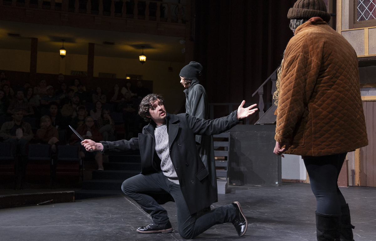 An actor crouches on a stage and gestures at another actor facing away from the camera. Audience members can be seen seated beyond the stage.