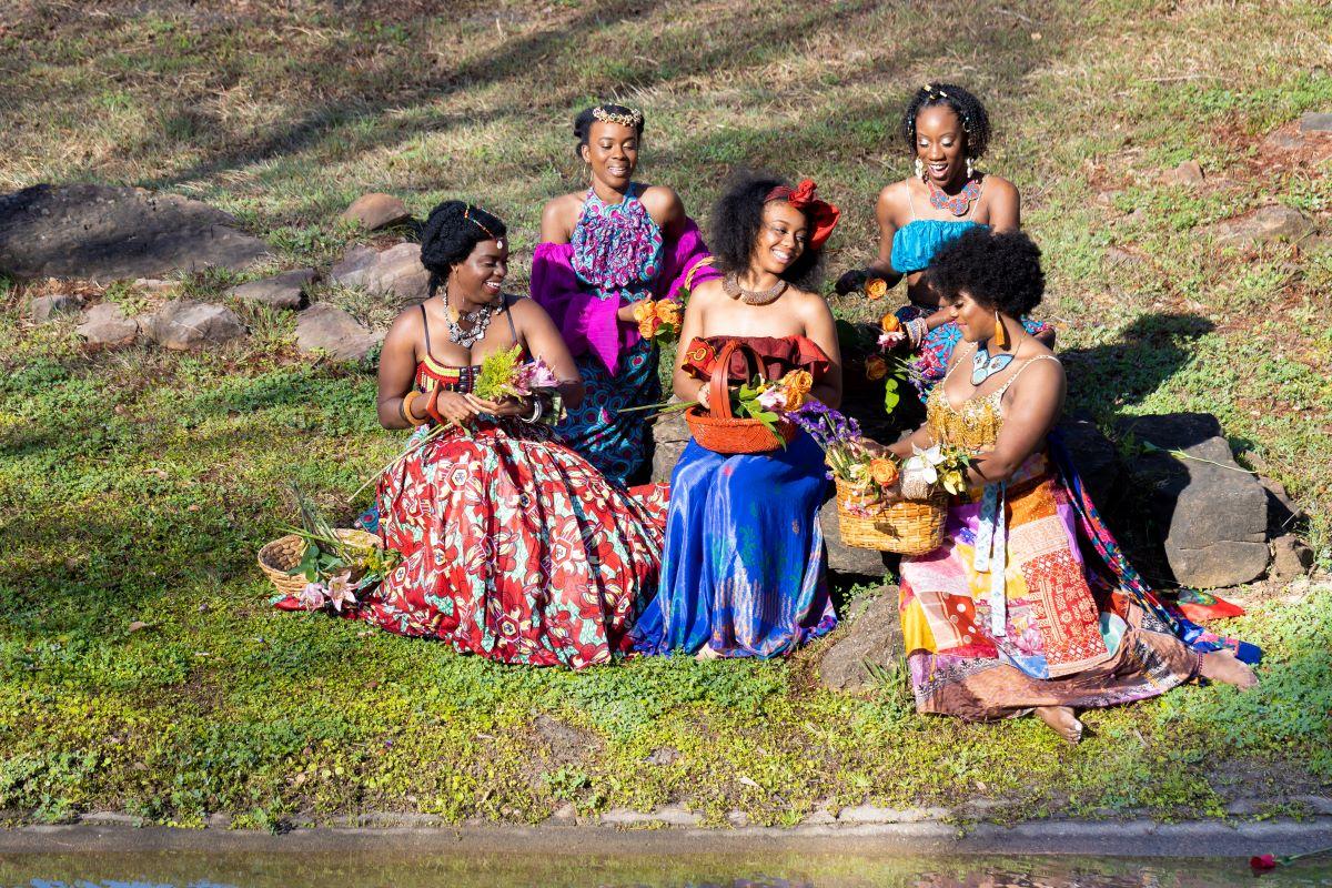 A group of five women in vibrant dresses sit on grass.