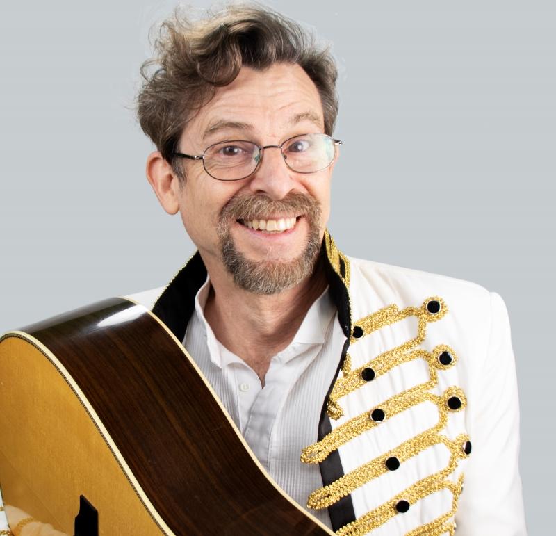 Joe McDermott smiles playfully at the camera while holding his guitar in one corner of the photo. He wears a marching band jacket and a white button-up shirt.