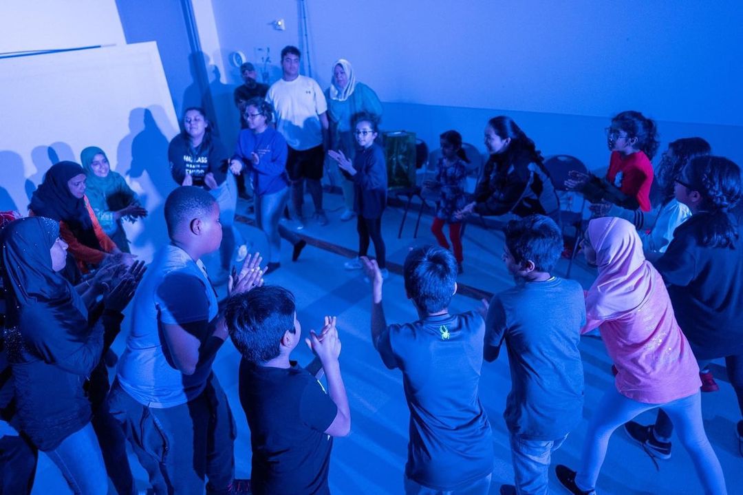 A circle of about 20 students stand and participate in an activity led by Brave Little Company. Their hands are raised as they clap, and they lean into the circle, towards each other. Photo by Alan Nguyen.