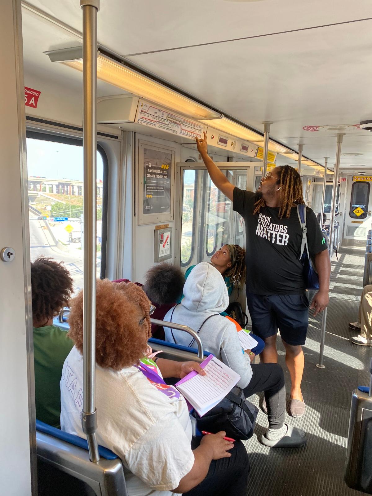 An instructor stands in a light rail car and points at a route map for students.