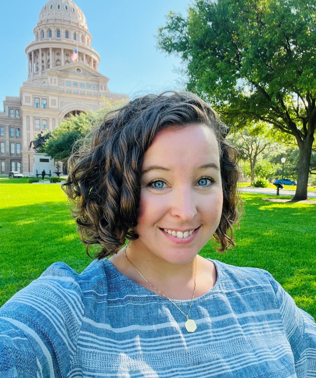 Spencer Zamora grins in front of the Texas Capitol building. She has blue eyes and curly brown hair. Spencer wears a blue-and-white patterned top.