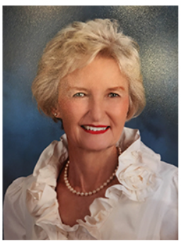 A headshot of TCA Commissioner Patty Nuss. Her short blonde hair is feathered and she smiles at the camera. She wears a ruffled white shirt and a strand of pearls.