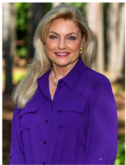 A headshot of TCA Commissioner Nancy C. Windham. She smiles warmly. Her long blonde hair drapes across one shoulder. She wears a bright blue button-up shirt and stands in front of some trees.