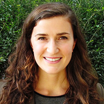 A headshot of Katy Sauer. She looks straight at the camera and smiles. Her wavy brown hair flows over both shoulders. She wears a grey blouse with black trim, and stands in front of a leafy green background.