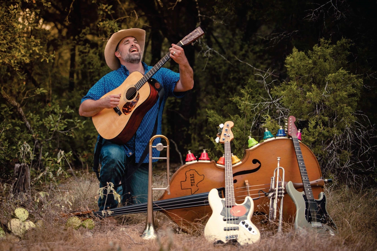 Will Dupuy sits outside strumming his guitar, with other instruments nearby.