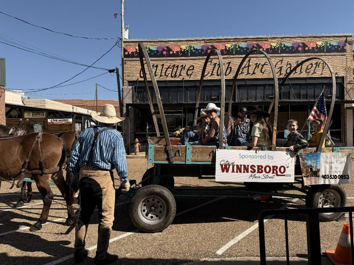 A man in western wear stands in front of a wagon full of people