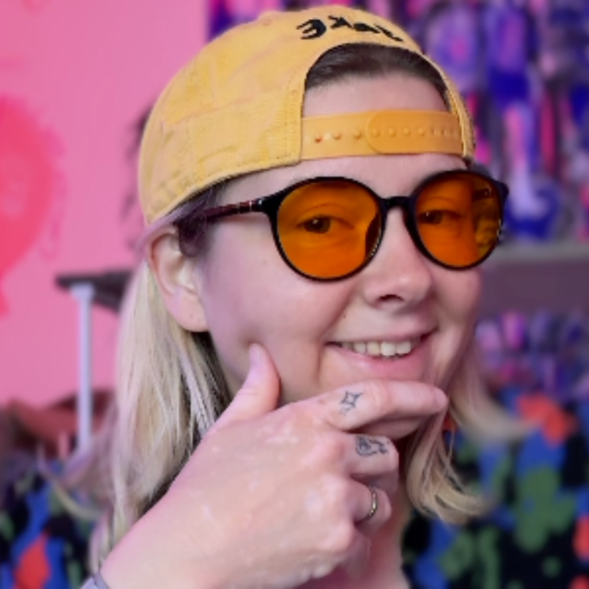 Casey Chavez holds her hand to her chin and smiles in front of a colorful background. She wears a yellow ballcap backwards and tinted glasses.