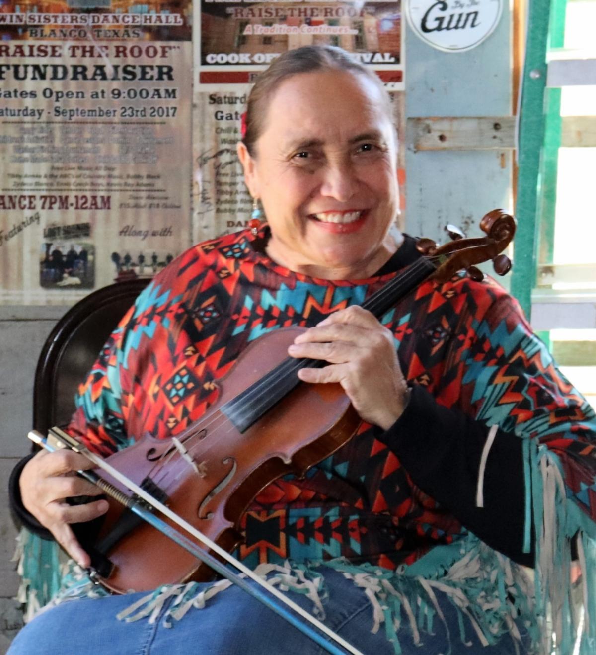 Belen Escobedo is seated and holding her fiddle across her chest as she smiles at the camera.