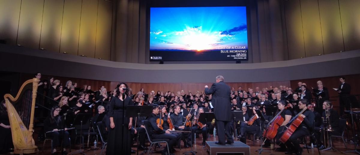 Bay Area Chorus performs with a featured soloist and musicians on stage. A screen on the wall shows a picture of the sun rising in a blue sky.