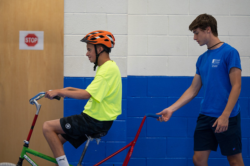 A staff member walks behind a person riding a bike with a handle attached to the back.