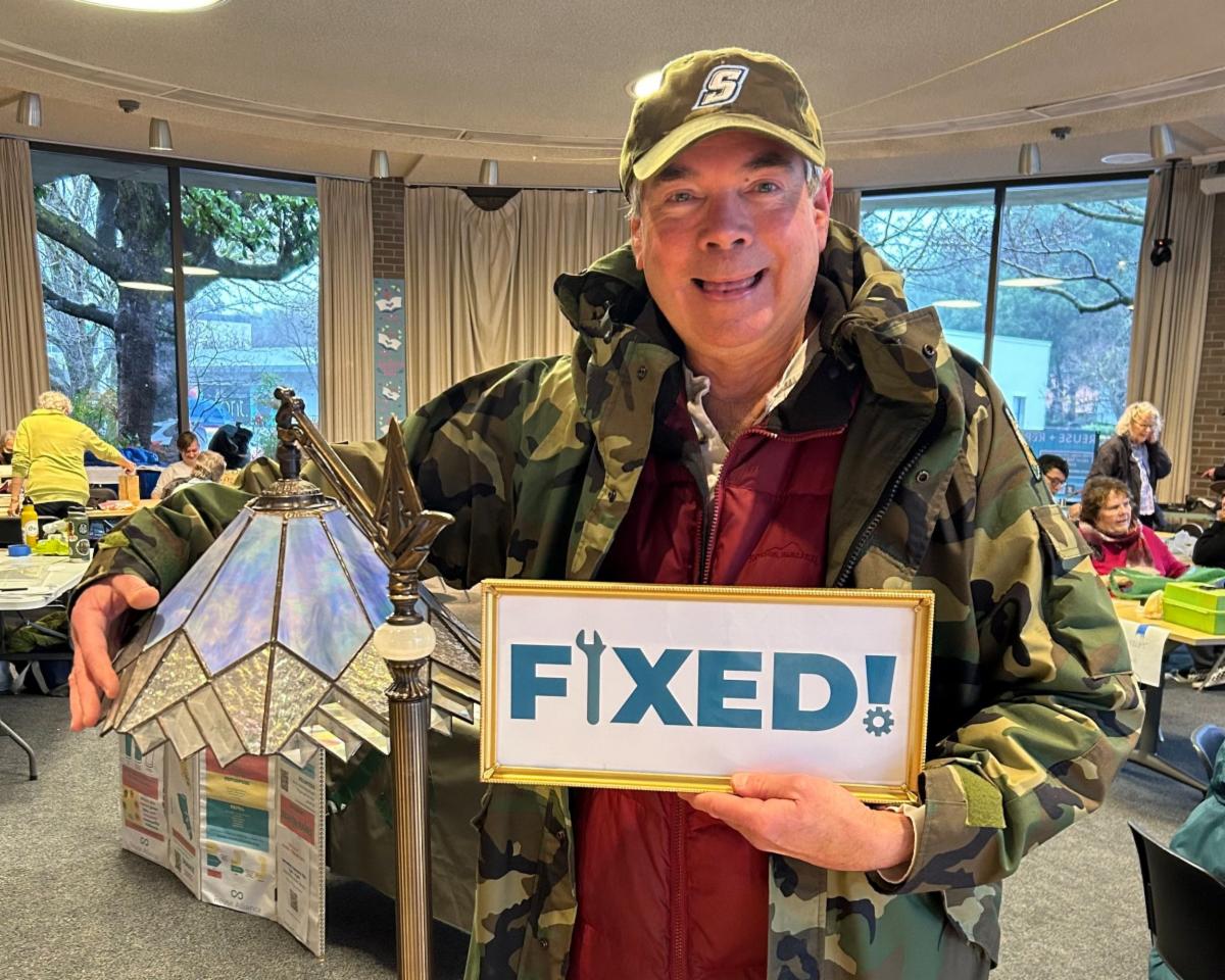  A smiling man wearing a camouflage jacket and cap holds a restored stained-glass lamp in one hand and a framed sign that reads FIXED in the other
