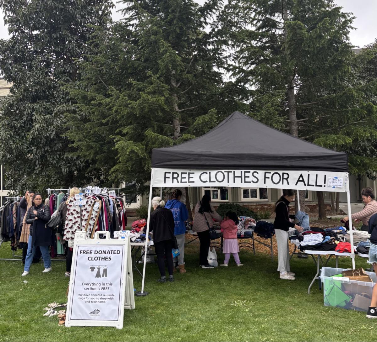 clothing swap event outdoors with a large black tent labeled FREE CLOTHES FOR ALL! Volunteers and visitors browse racks and tables filled with donated clothing.