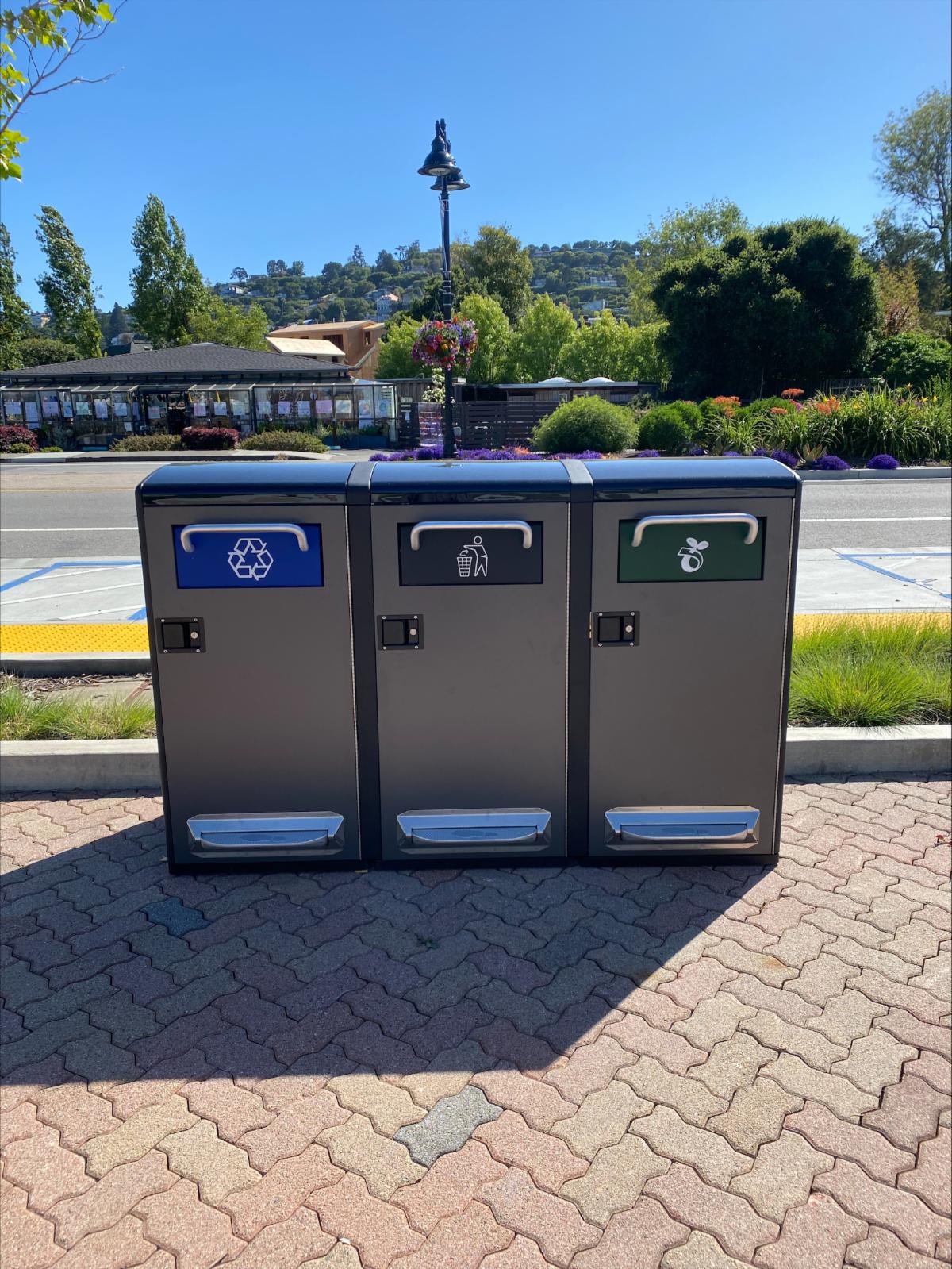 three new waste receptacles for landfill recycling and compost sit next to the street in front of the library