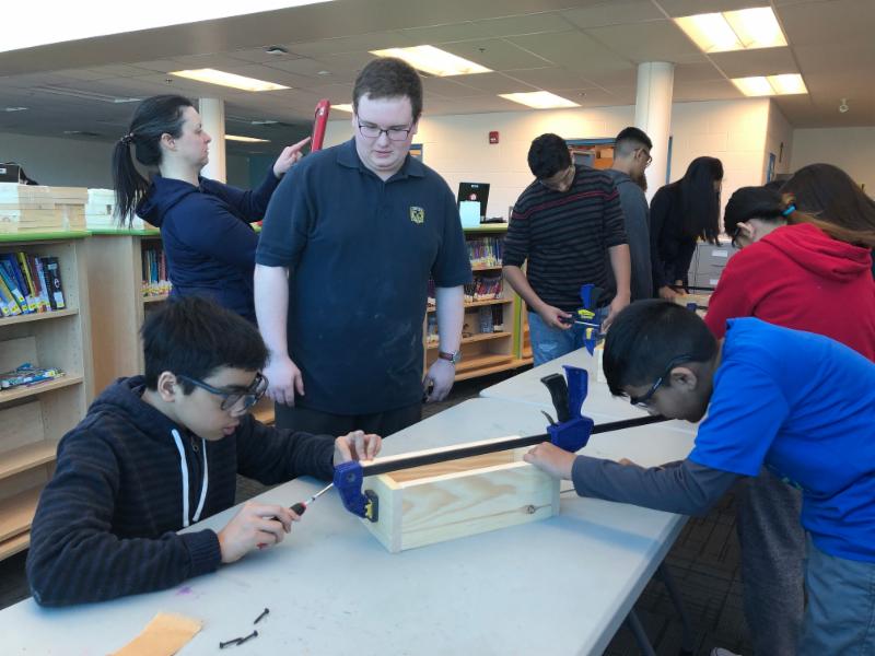 Male secondary student supporting two male students as they assemble a wooden box.