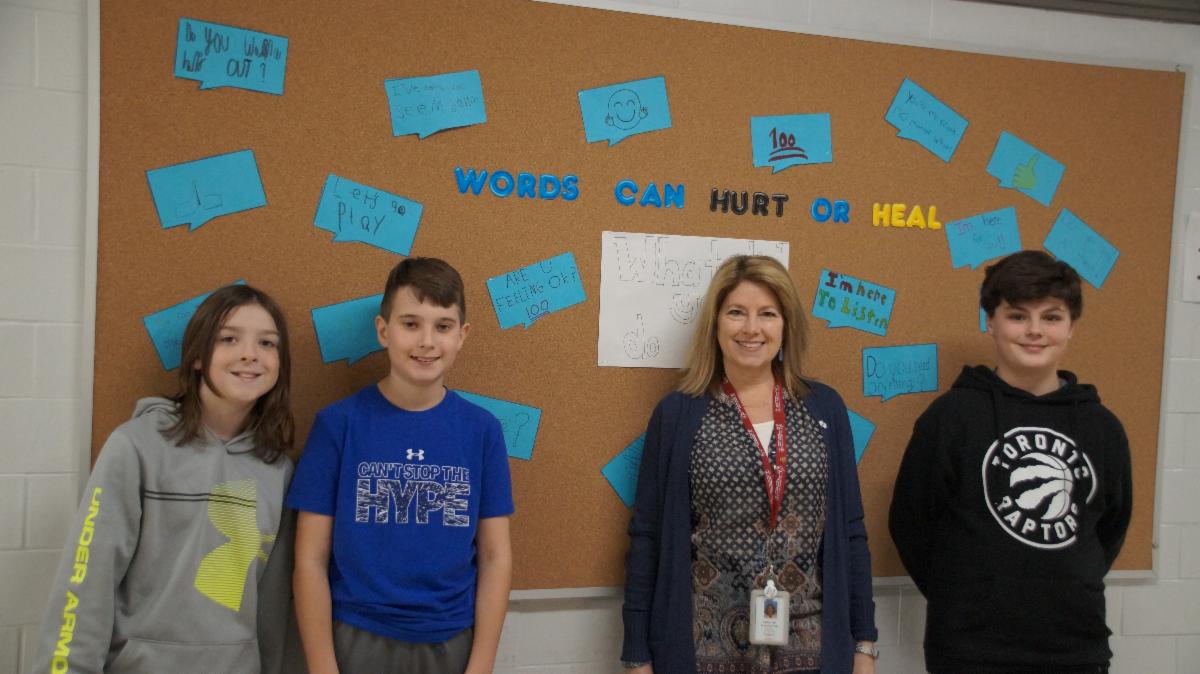 Three male students with female adult standing against bulletin board with words that can heal mental health