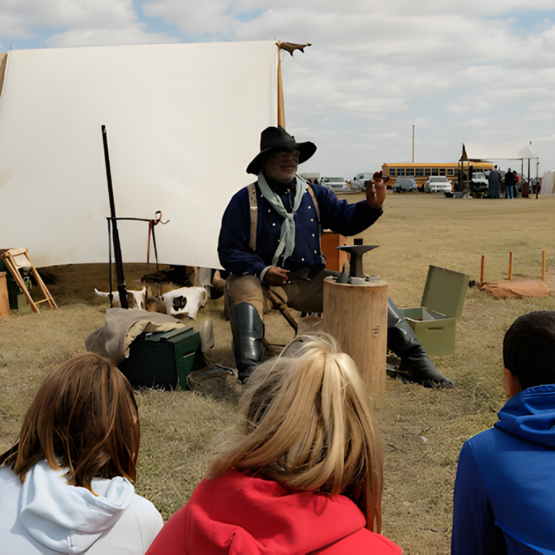 Back of three children watching a man in costume tell a story in front of a covered wagon
