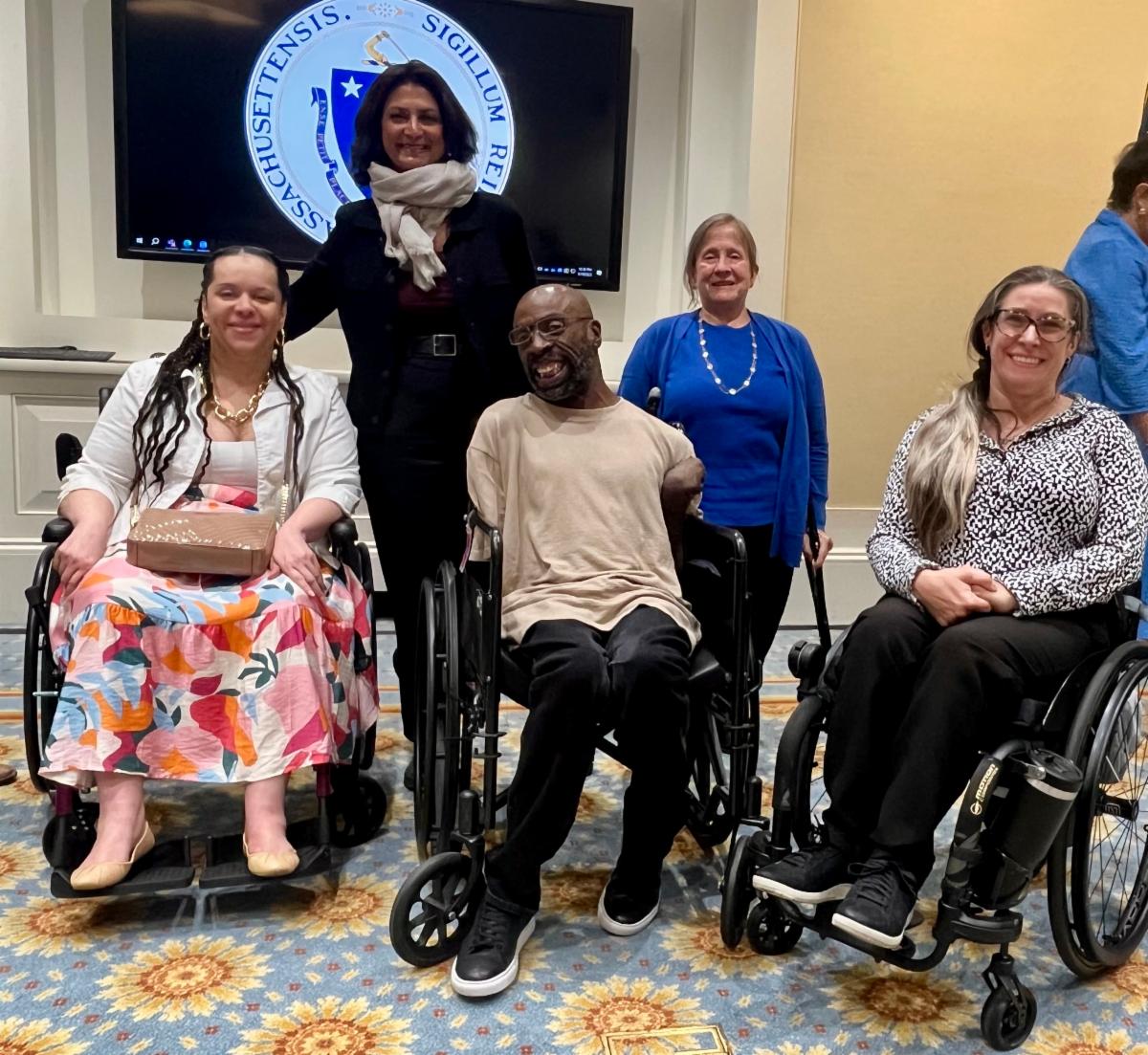 From left to right, Heather Watkins, Dr. Monika Mitra, Keith Jones, Linda Long-Bellil, and Nicole Lomerson pose together and smile for a group photo indoors. Heather Watkins is wearing a white jacket and a colorful floral skirt, Dr. Mitra is wearing a black suit with a red shirt and a beige scarf, Keith Jones is wearing a beige shirt and black trousers, Linda Long-Bellil is wearing a blue cardigan on a blue shirt, and Nicole Lomerson is wearing a black-and-white patterned top. The Massachusetts state seal is visible on a screen behind them, and the room has a blue and yellow floral carpet and cream-colored walls.