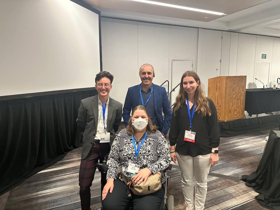 Group photo from the conference featuring (from left to right): Alex Levine, Laurin Bixby, Ilhom Akobirshoev, and Victoria Lajous. Alex is wearing glasses, a white shirt, and a blazer. Laurin, seated in a wheelchair, is wearing a black-and-white top and a face mask. Ilhom is dressed in a blue blazer, and Tori is in a black blouse and white pants. Behind them is a formal conference setup with a projector screen, podium, and rows of chairs.