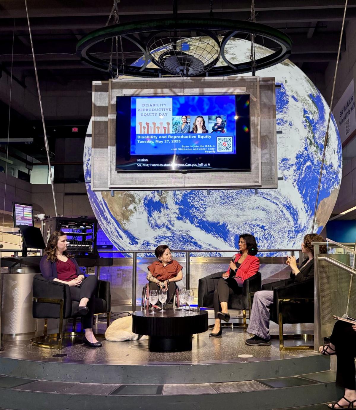 Panelists (from L-R: Tiffany L. Panko, Mia Ives-Rublee, and Monika Mitra) are seated in a semi-circle together with the presenter on a stage, engaged in a panel discussion. Behind them is a large digital screen that projects the event flyer. Behind the screen is a backdrop of a massive image of Earth.