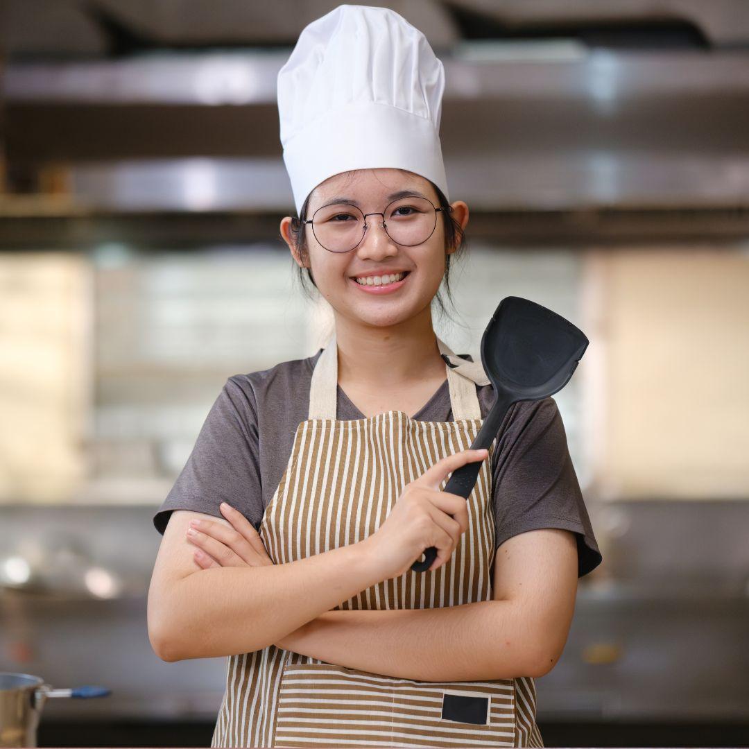 Teen in a kitchen with a chef's hat and apron, holding a spatula.