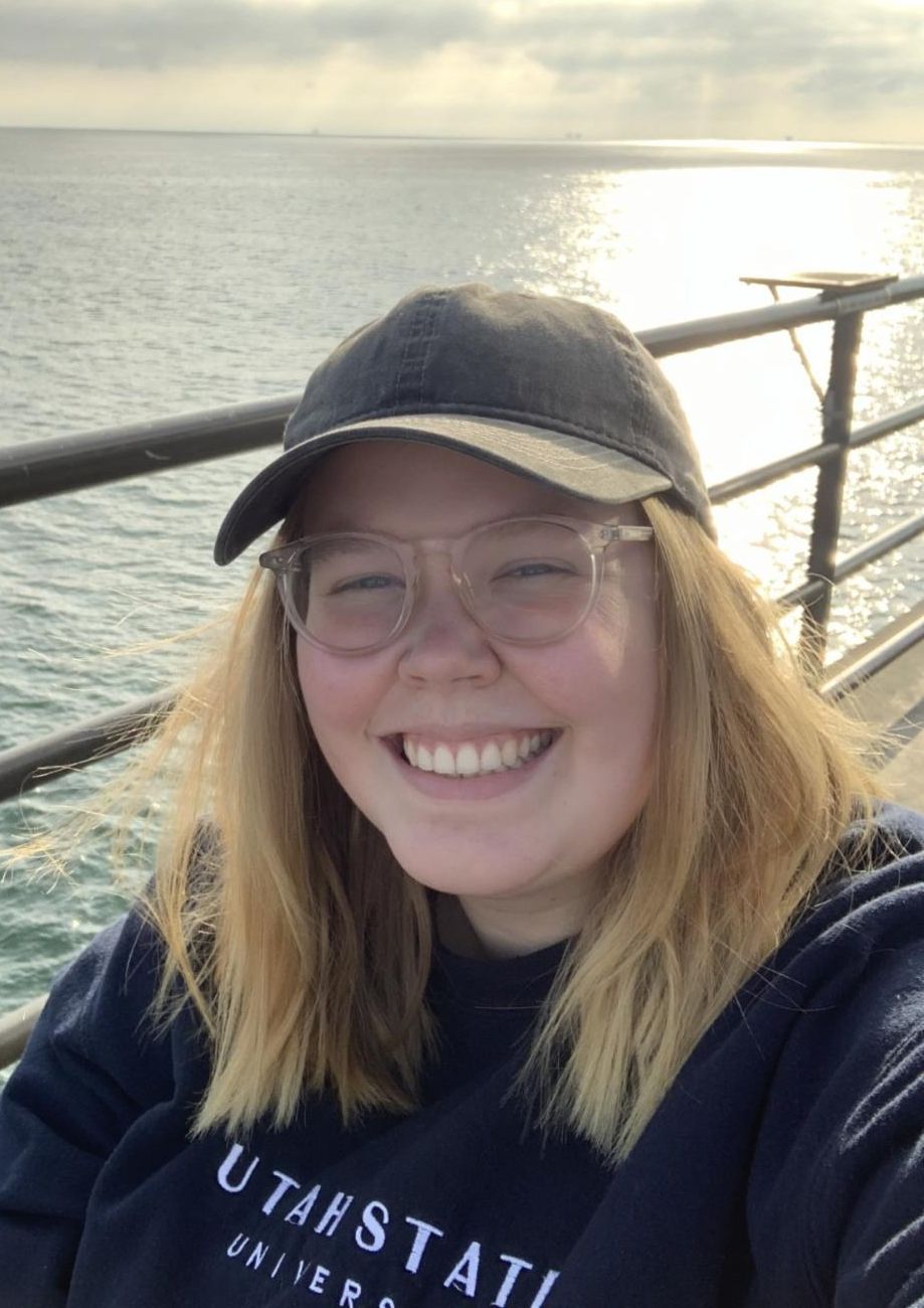 Hannah smiling in front of ocean wearing Utah State sweatshirt.
