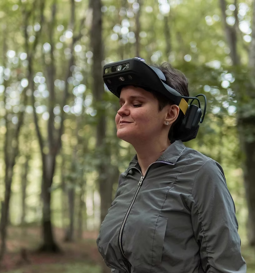 A woman stands in the woods wearing Lumen glasses, a black device that wraps around her forehead and the back of her neck.
