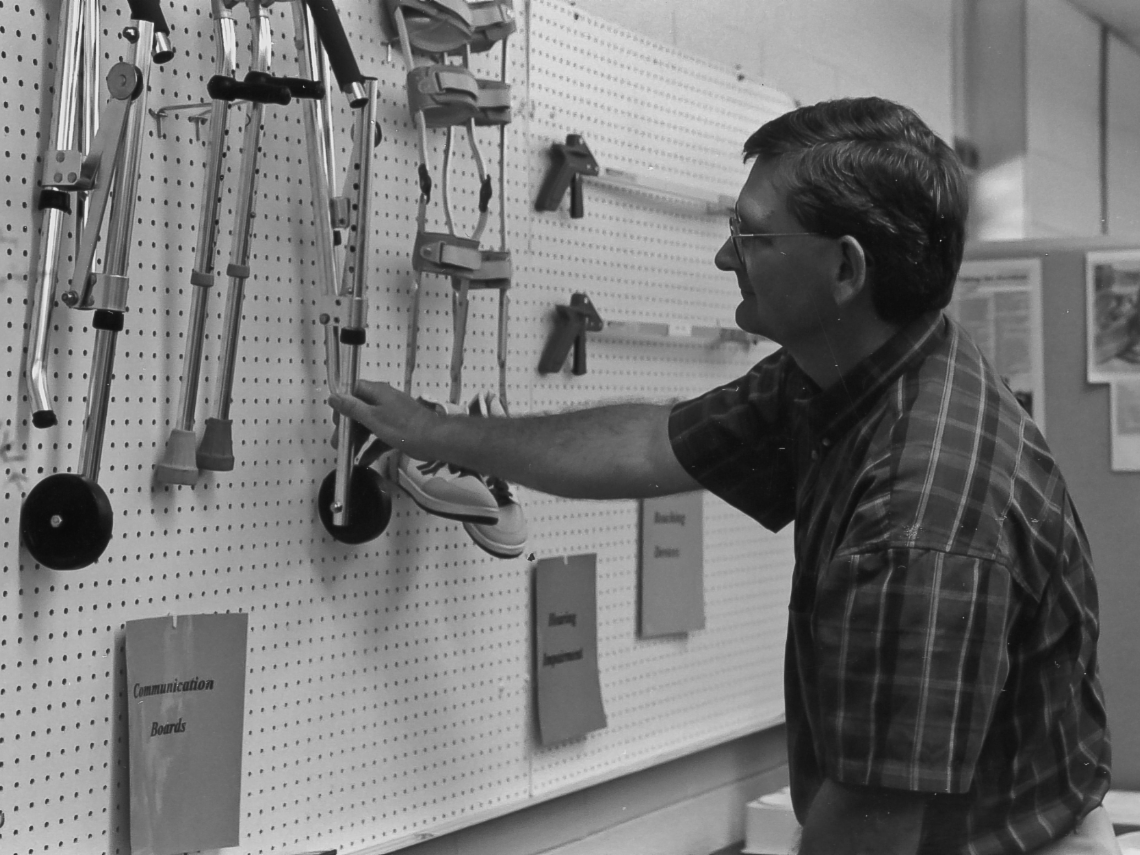 Black and white photo of man sorting through wall of assistive technology.